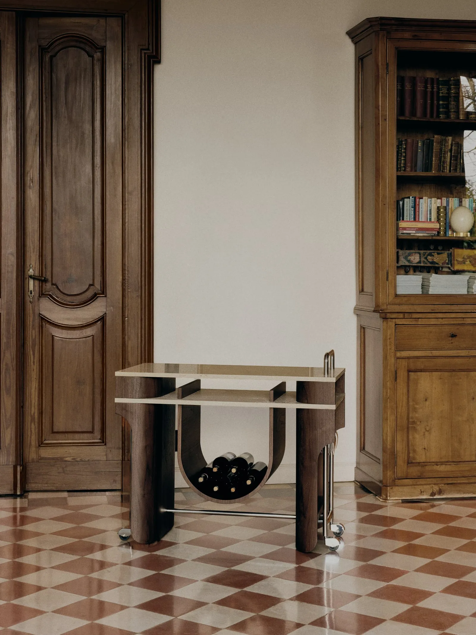Arcus Bar Cart with wine bottles in its curved cradle, beside a carved door and a wooden bookcase