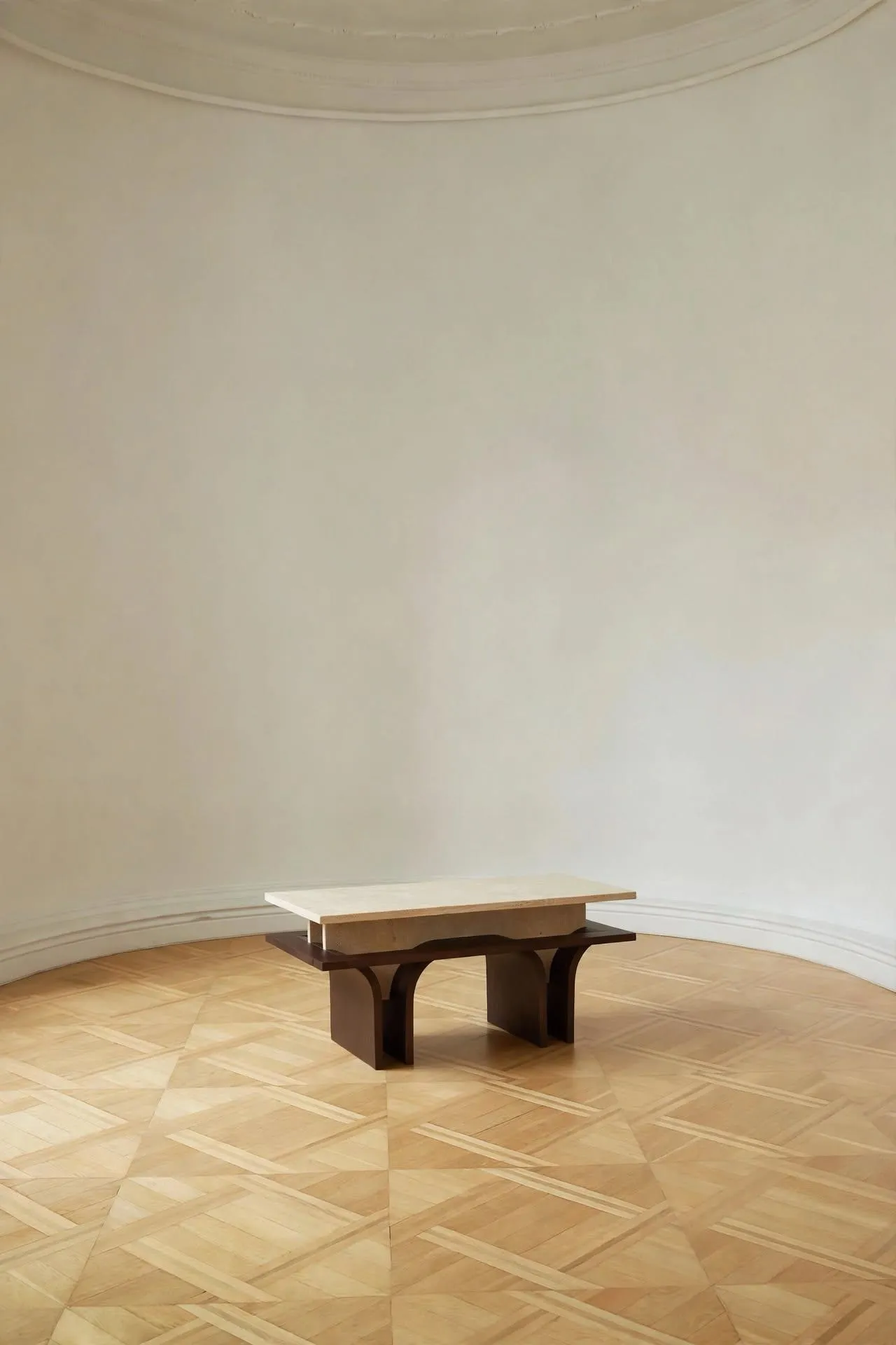 Arcus bench with a pale stone top on arched dark wooden legs, in a round room with a parquet floor