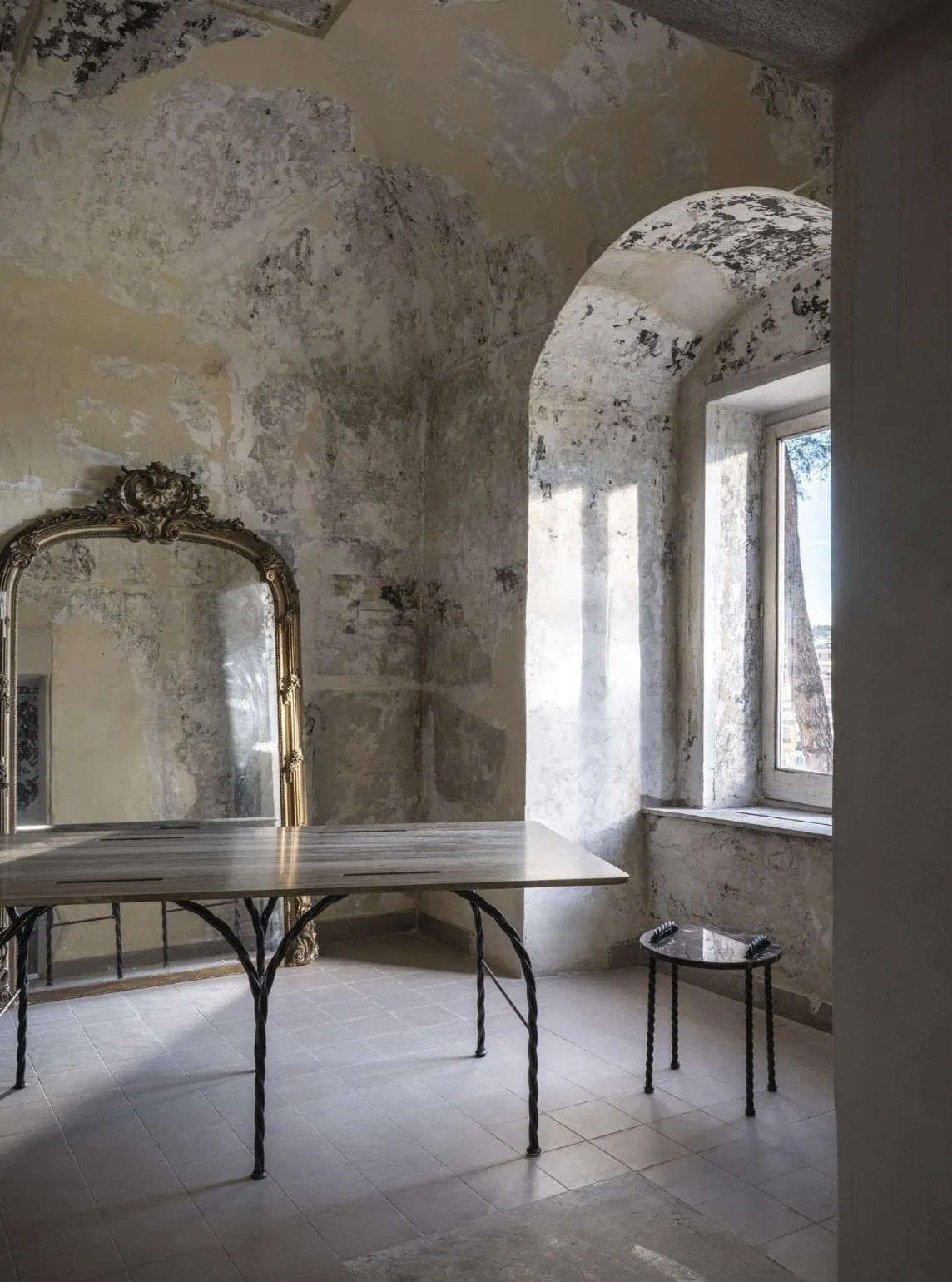 A long table and stool on twisted black metal legs in a room with distressed plaster walls and a gilt mirror