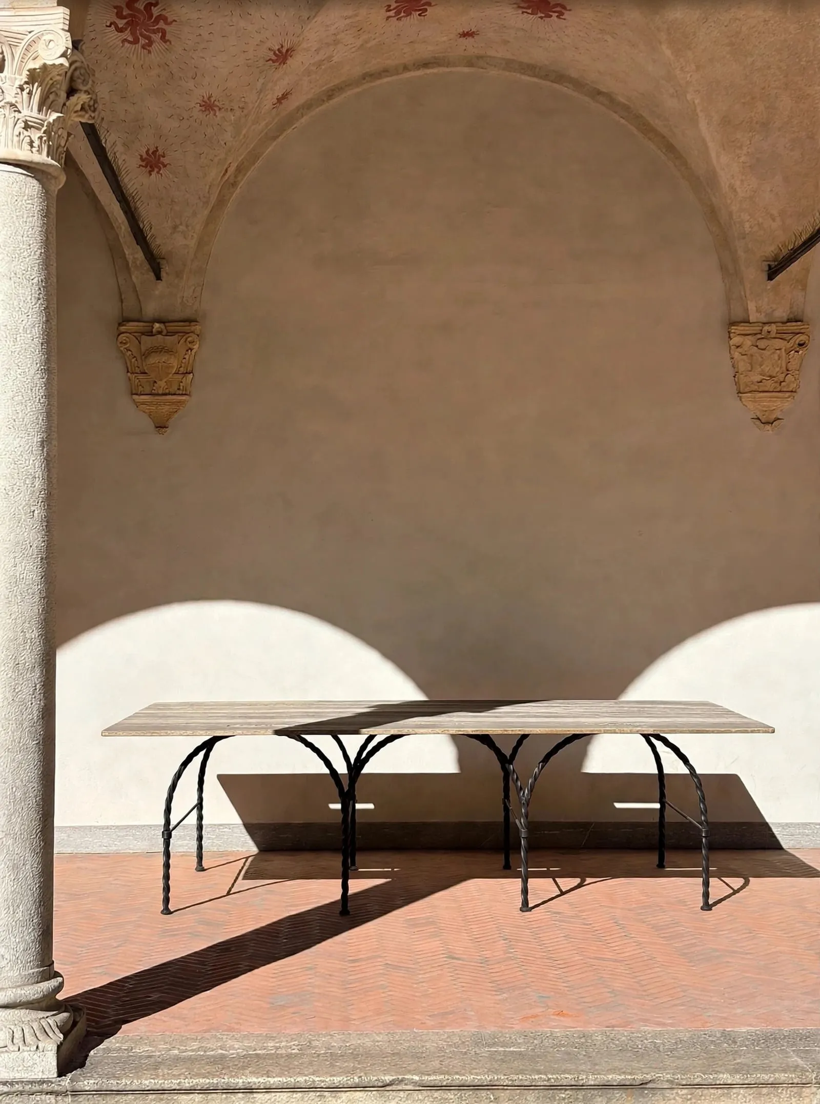 Tresse Città Table in a sunlit cloister, its arched legs against a curved wall