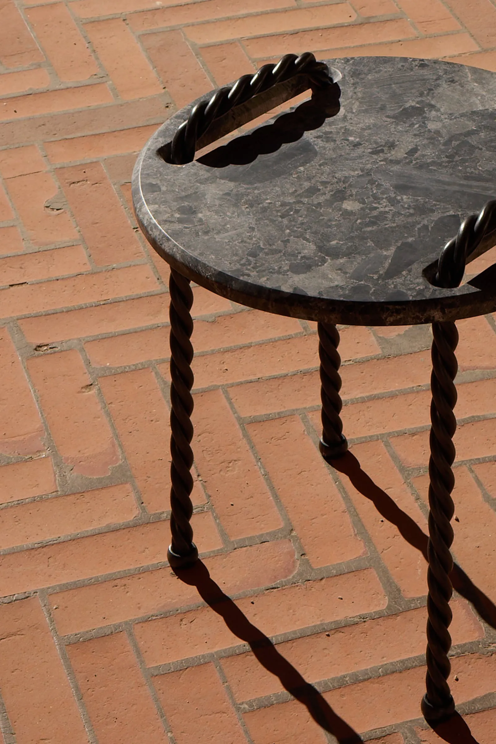 Close view of the Tresse Side Table: dark stone top and braided legs on herringbone bricks