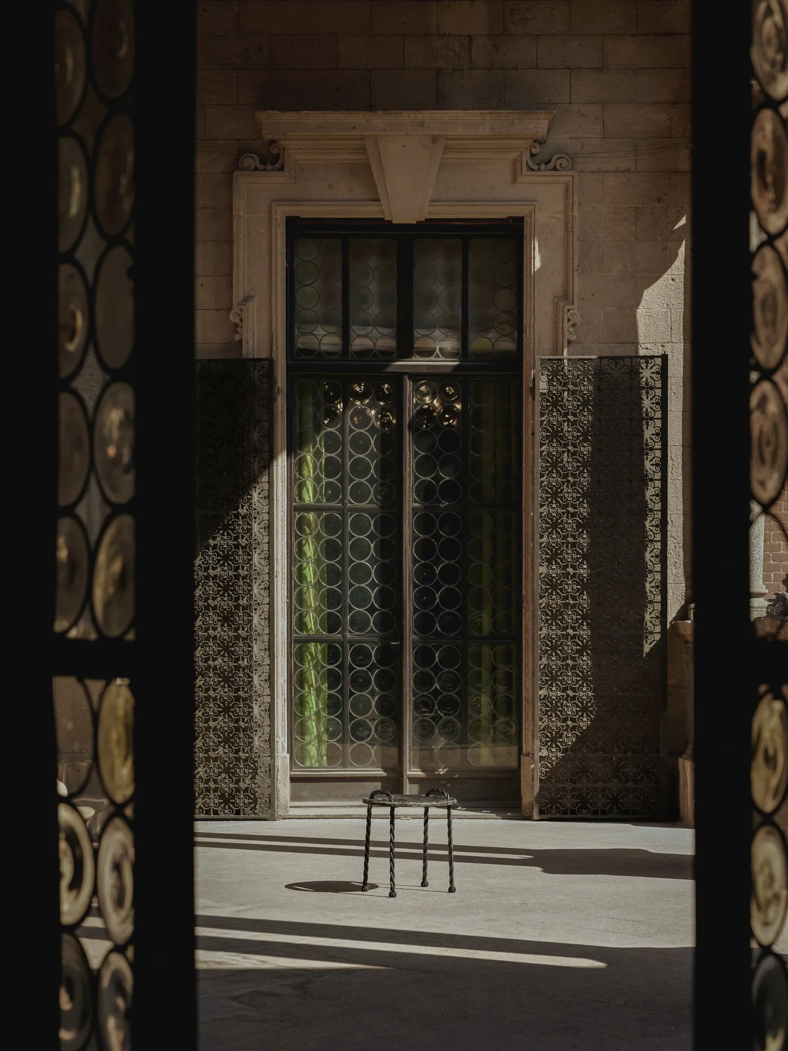 Tresse Side Table seen through a doorway of round leaded-glass panes, in a sunlit stone courtyard